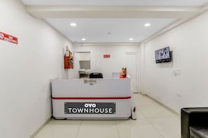 Una zona de recepció amb un taulell de recepció amb la indicació 'OYO TOWNHOUSE', un cartell de 'NO FUMAR', una càmera de seguretat i una televisió penjada a la paret.