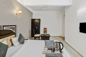 Una habitació d'hotel amb llit, taula de nit, escriptori, cadira, televisió i ventilador de sostre.