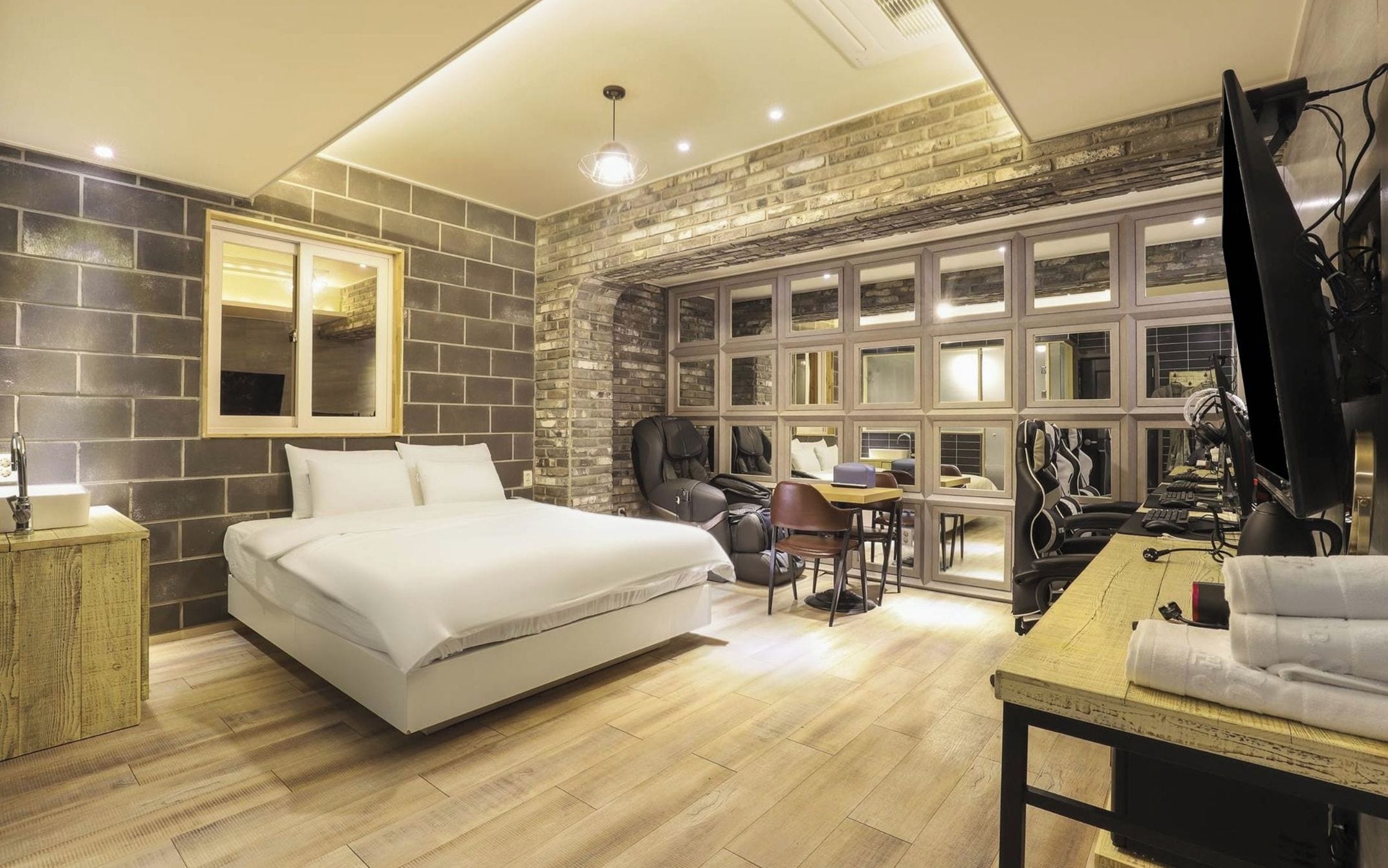 A modern hotel room with a large bed, a massage chair, a desk with a computer, and a large window with multiple panes.