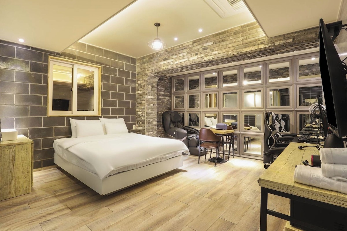 A modern hotel room with a large bed, a massage chair, a desk with a computer, and a large window with multiple panes.