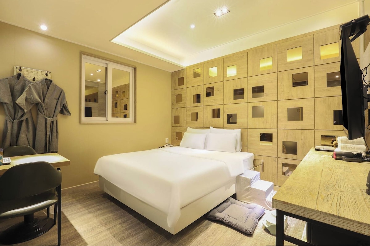 A hotel room with a large bed, a desk with a chair, a mirror, and a wardrobe with robes.