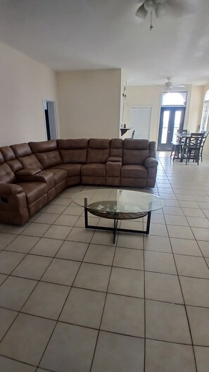 Una sala de estar con un sofá seccional marrón, una mesa de centro de vidrio, y un comedor con sillas y una mesa al fondo.