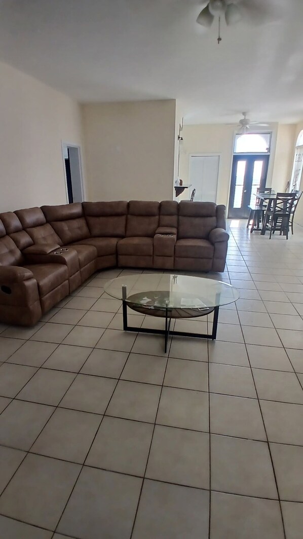 A living room with a brown sectional sofa, a glass coffee table, and a dining area with chairs and a table in the background.