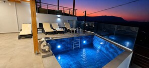 Una piscina sul tetto con poltrone prendisole, ringhiera in vetro e vista sul tramonto.