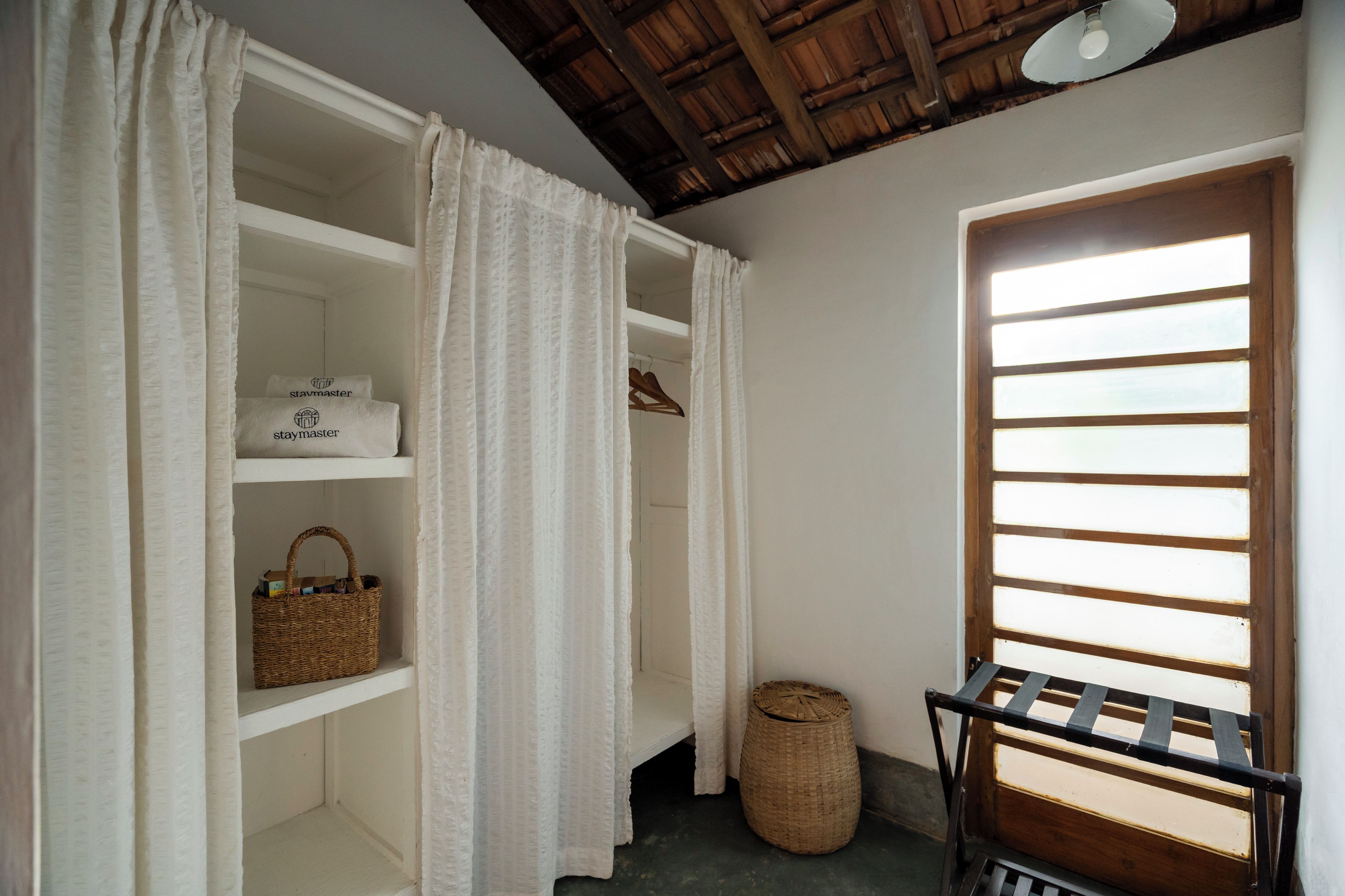 A room with a wooden ceiling, a window with horizontal blinds, a wicker basket on a shelf, and a small table with a metal frame.