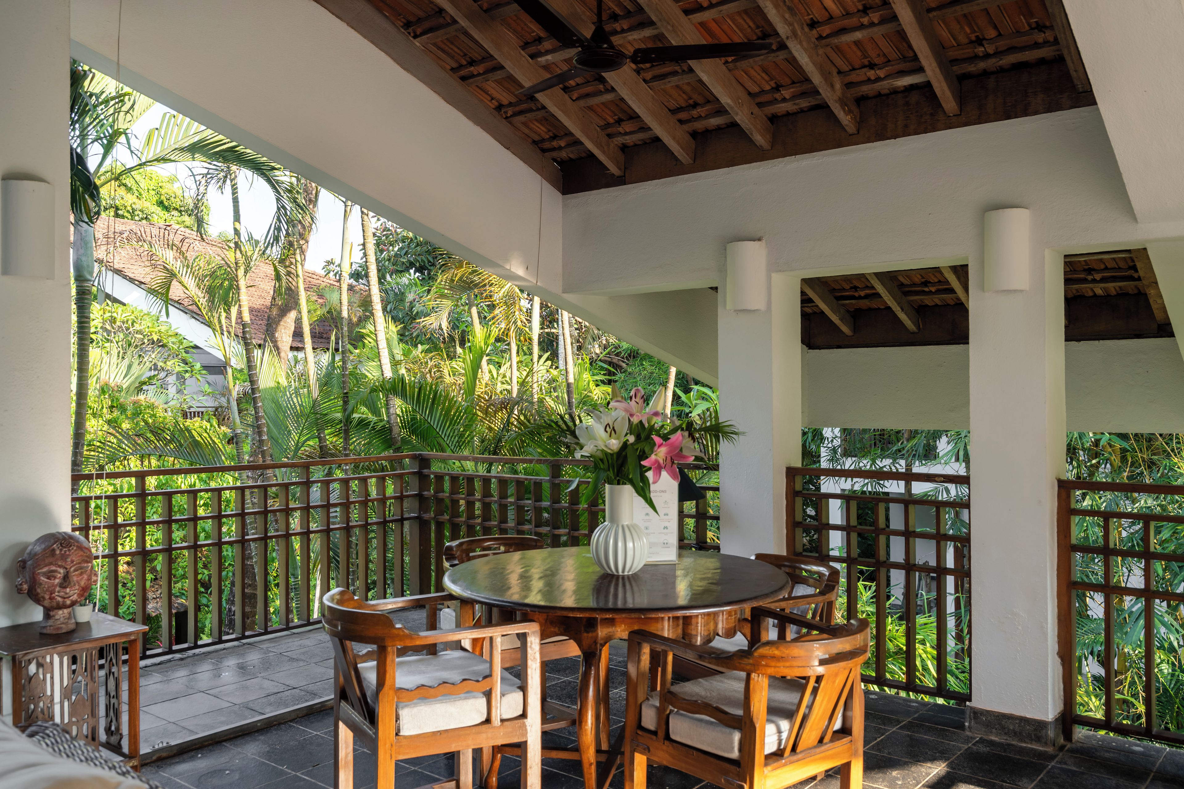 A covered outdoor seating area with a round table and chairs, surrounded by a wooden fence and lush greenery.