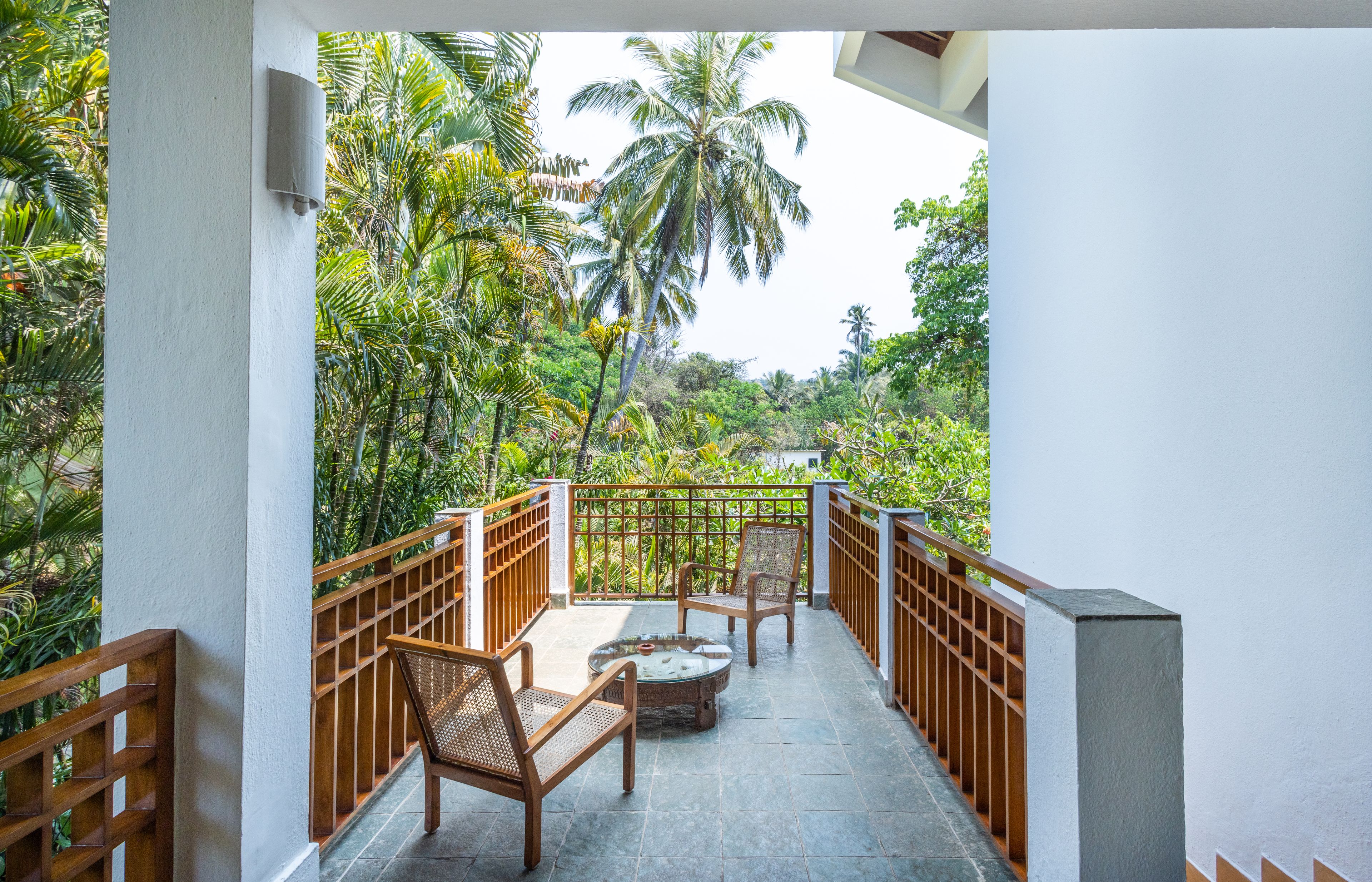 A balcony with wooden railings, a table, and two chairs, overlooking a lush garden with palm trees.