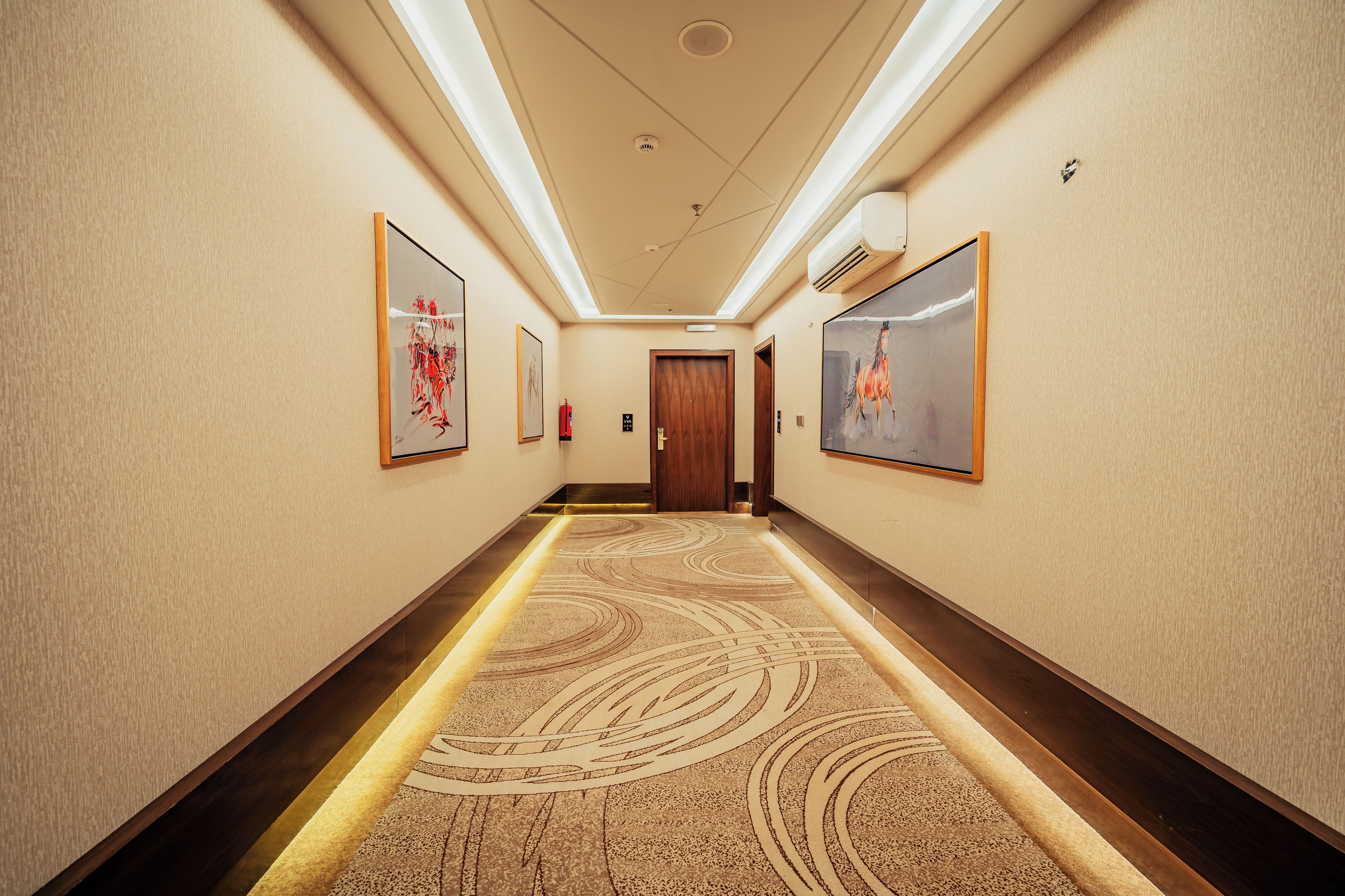 A hotel hallway with a patterned carpet, framed artwork on the walls, and a wooden door at the end.
