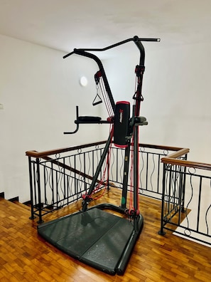 A stationary exercise bike with a foldable platform, located in a room with wooden flooring and a metal railing.