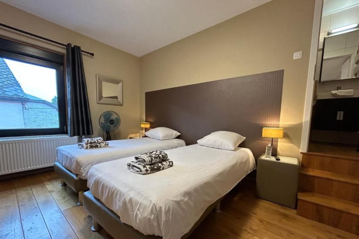 A hotel room with two beds, a window with curtains, a fan, a nightstand with a lamp, and a staircase.