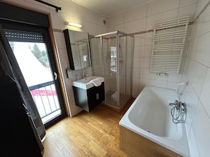 A bathroom with a glass-enclosed shower, a white bathtub, a sink with a mirror, and a door leading to an outdoor area. - Race & Rooms (Stavelot)