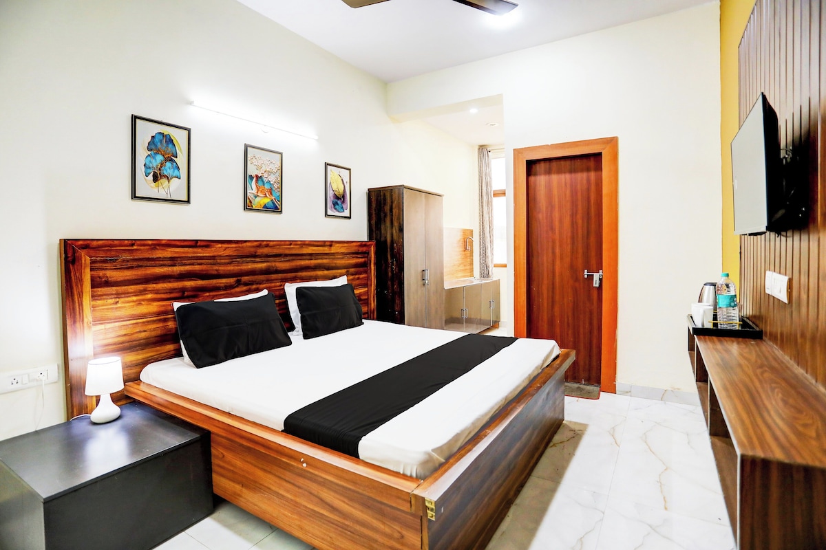 A hotel room with a wooden headboard, a bed with black and white bedding, a bedside table with a lamp, a wardrobe, a bathroom with a shower, and a TV mounted on the wall.