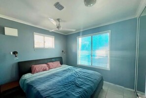 Une chambre à coucher avec un lit, deux lampes fixées au mur, un ventilateur de plafond et une fenêtre avec des stores.