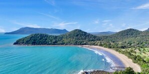 Un paysage côtier avec une plage, des collines boisées et un ciel dégagé.