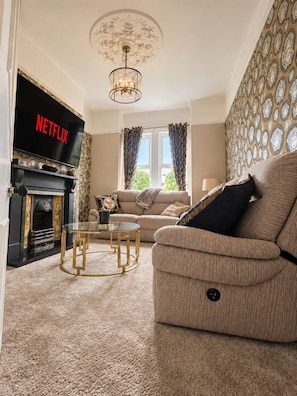 Un salon avec une cheminée, un canapé, une table basse en verre et un téléviseur affichant le logo  « NETFLIX ».