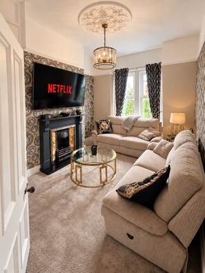 En stue med et fjernsyn, der viser 'NETFLIX', en sofa, et sofabord i glas, en pejs og tapet med mønster.