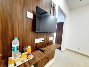 Habitació amb televisió penjada, panells de fusta, paret blanca, una porta i una taula petita amb una ampolla i un got.