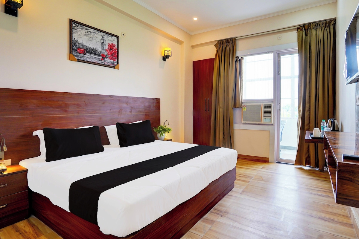 A hotel room with a large bed, a wooden headboard, a painting on the wall, a desk with a chair, and a window with curtains.