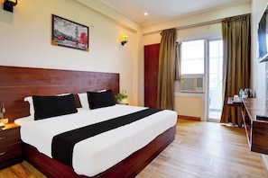 A hotel room with a large bed, a wooden headboard, a painting on the wall, a desk with a chair, and a window with curtains. - Super Hotel O by OYO THE VIRAAJ BANQUET (Greater Noida)