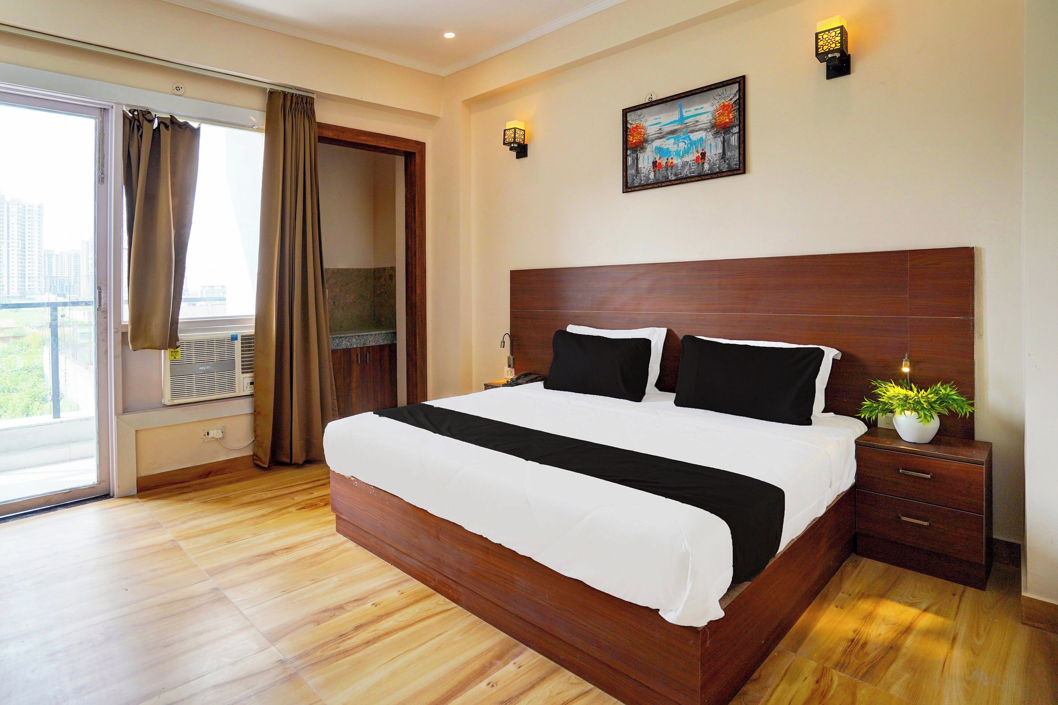 A hotel room with a large bed, a nightstand, a potted plant, and a cityscape view through the window.
