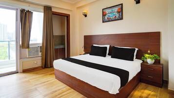 A hotel room with a large bed, a nightstand, a potted plant, and a cityscape view through the window.
