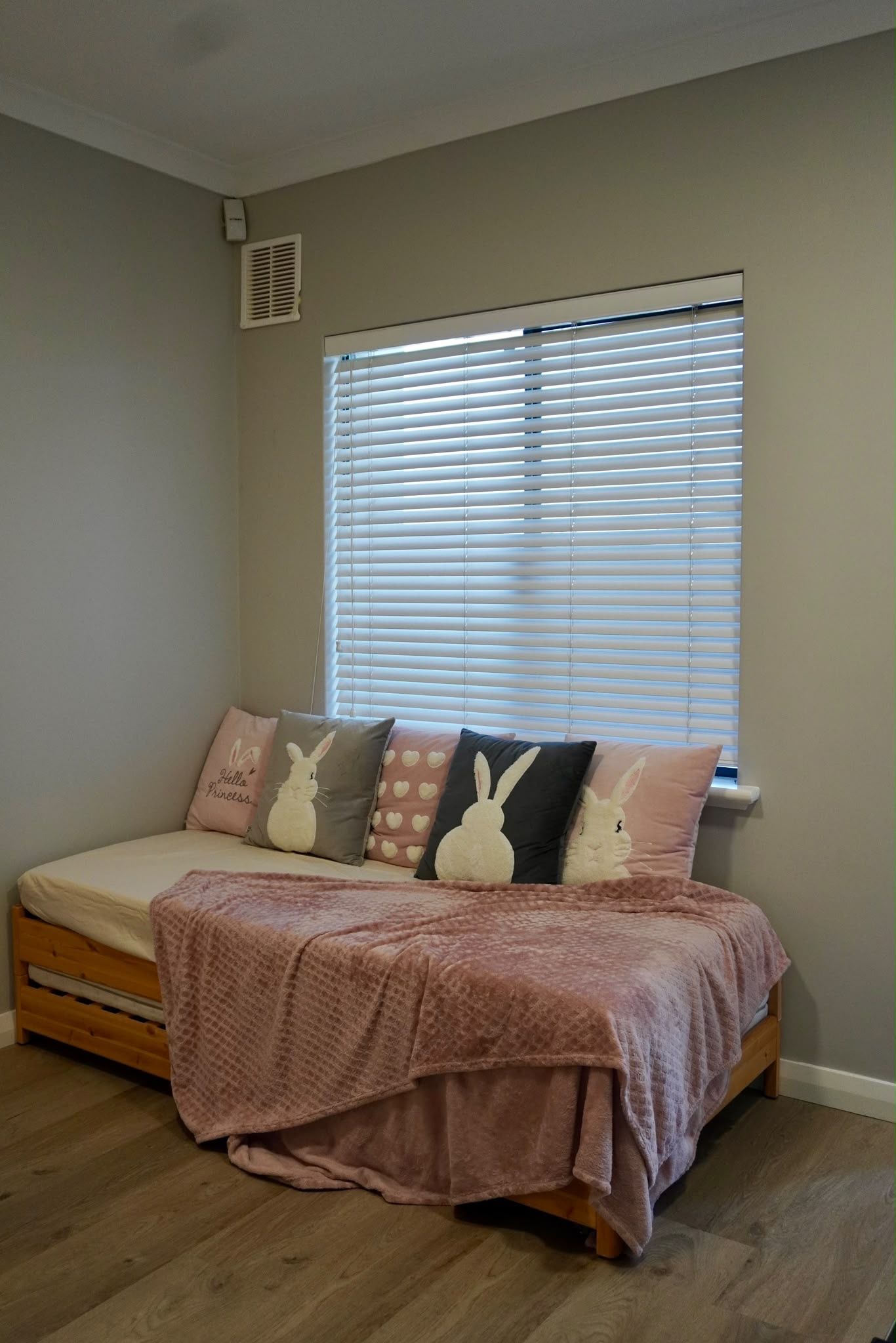 Une chambre avec un lit, une fenêtre avec des stores et une bouche d’aération fixée au mur.
