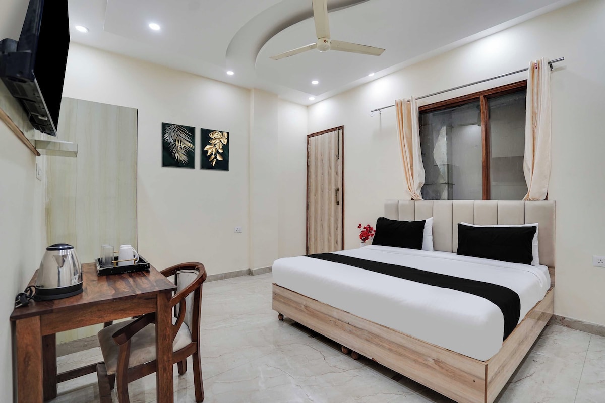 A hotel room with a bed, a desk with a kettle, a chair, a ceiling fan, and two wall art pieces.
