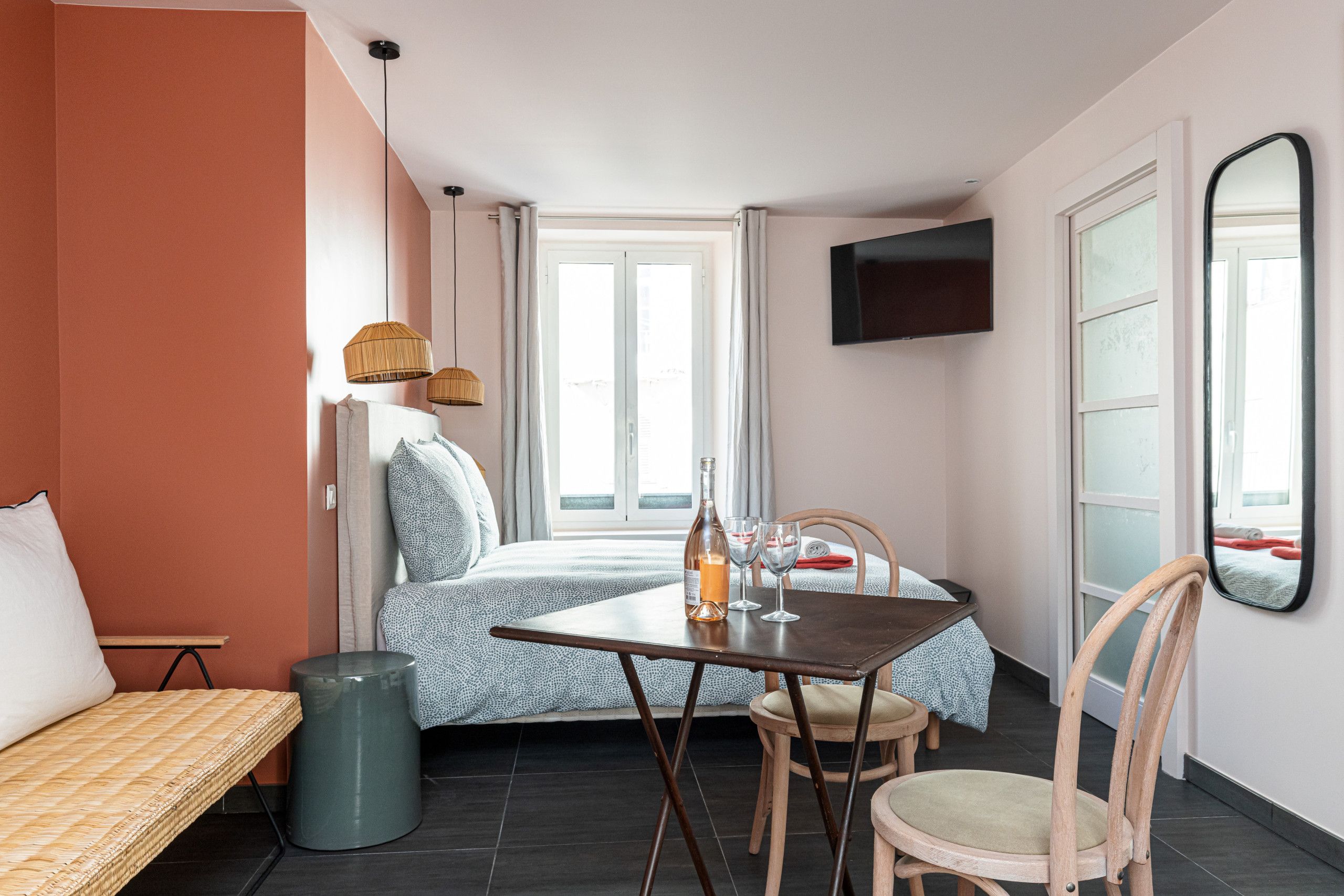 Een kleine hotelkamer met een bed, een tafeltje met een fles en glazen, een stoel, een prullenbak en een aan de muur bevestigde televisie.