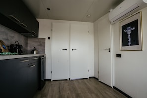 Cozinha moderna com máquina de lavar louça embutida, chaleira e máquina de café no balcão. Duas portas brancas com puxadores pretos, e um ar condicionado fixado na parede.