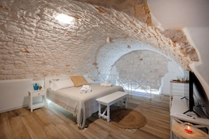 A bedroom with a stone ceiling, a bed, a bedside table, a nightstand, a lamp, a wooden floor, and a window. - Piazza Vecchia SUITE & SPA (Castellana Grotte)