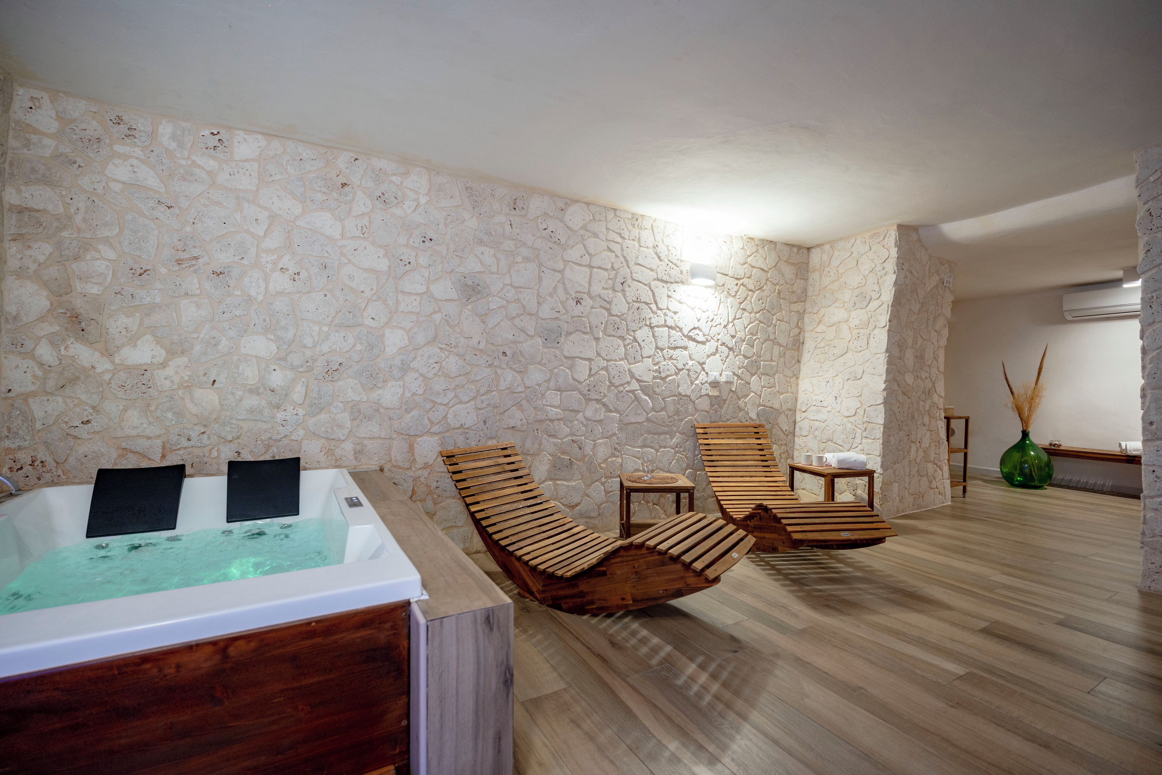Une salle de spa avec un mur en pierre, des chaises longues en bois, une table de massage et une petite table avec un verre.