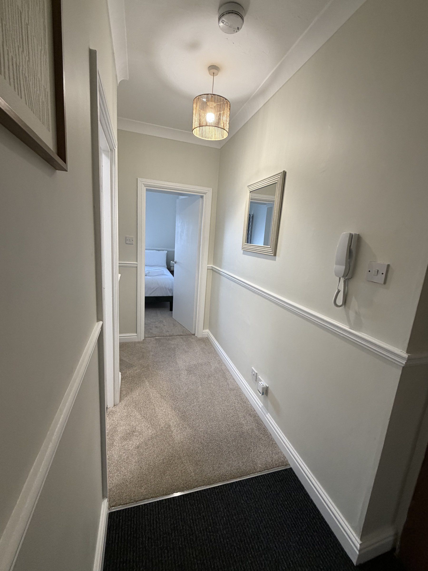 A hallway with a carpeted floor, a door leading to a bedroom, a wall-mounted telephone, and a hanging light fixture.