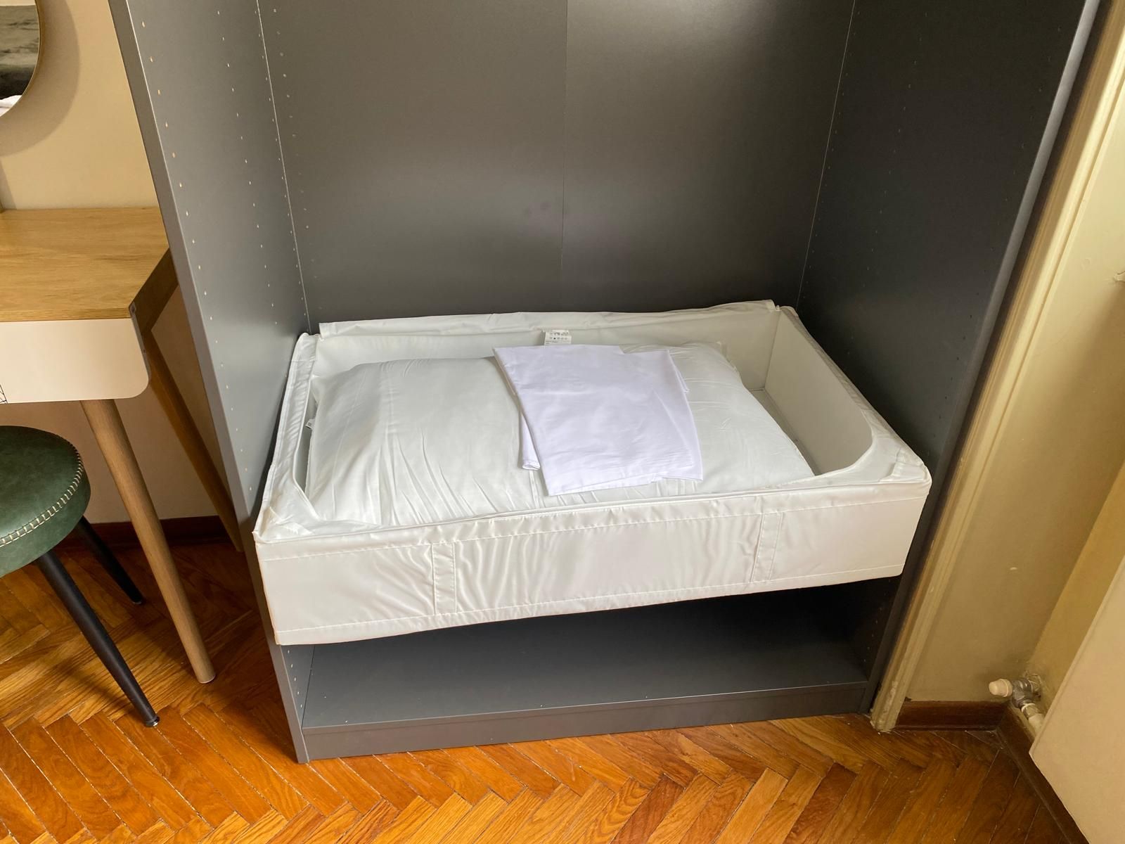 Ein kleines Bett mit einer weißen Matratze und einem zusammengeklappten weißen Handtuch, unter einer grauen Wand in einem Raum mit Holzboden.