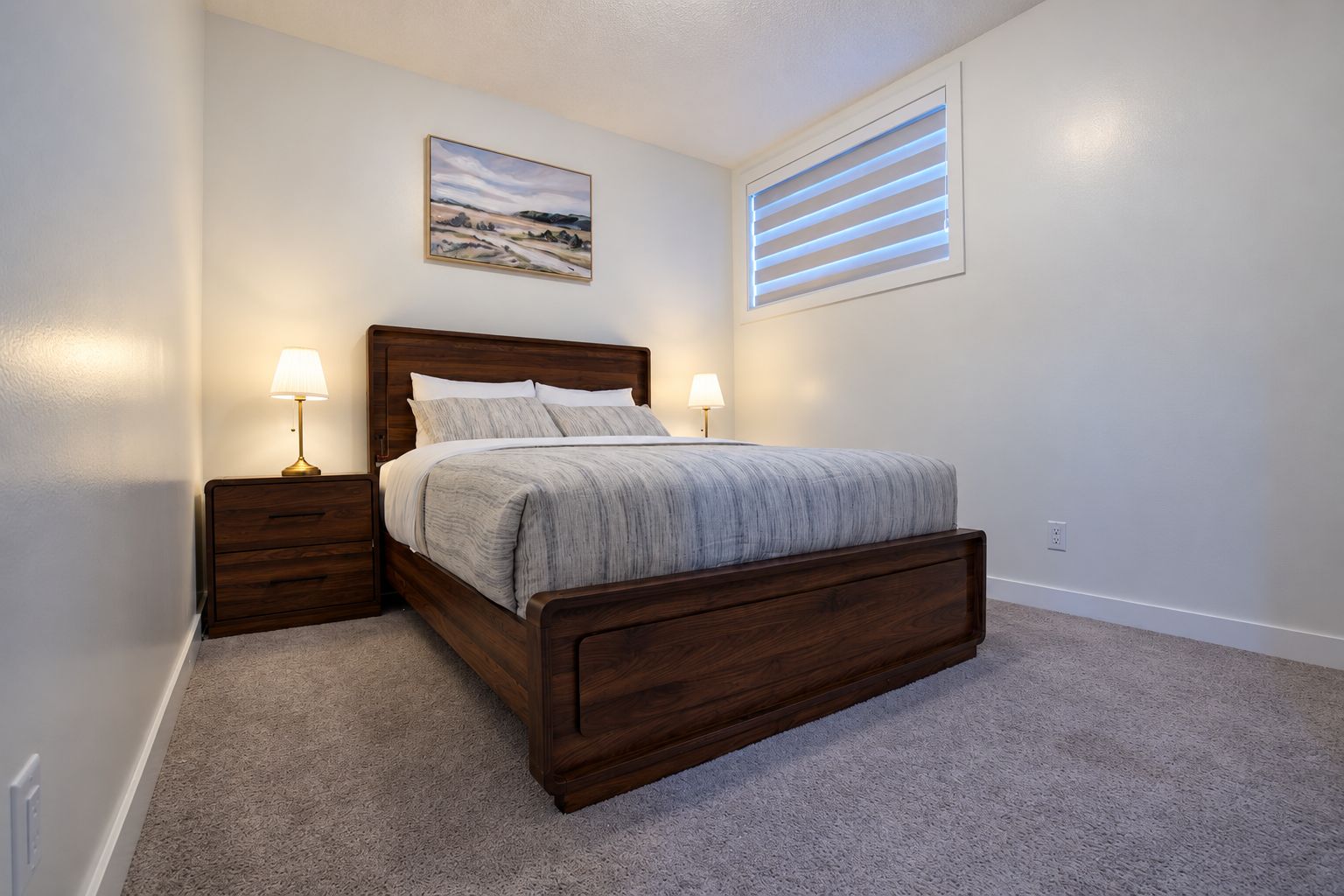 A bedroom with a wooden bed, two bedside tables with lamps, a carpeted floor, a painting on the wall, and a window with blinds.