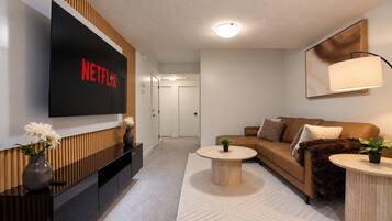A modern living room with a flat-screen TV displaying 'NETFLIX,' a sofa, two coffee tables, and a potted plant.