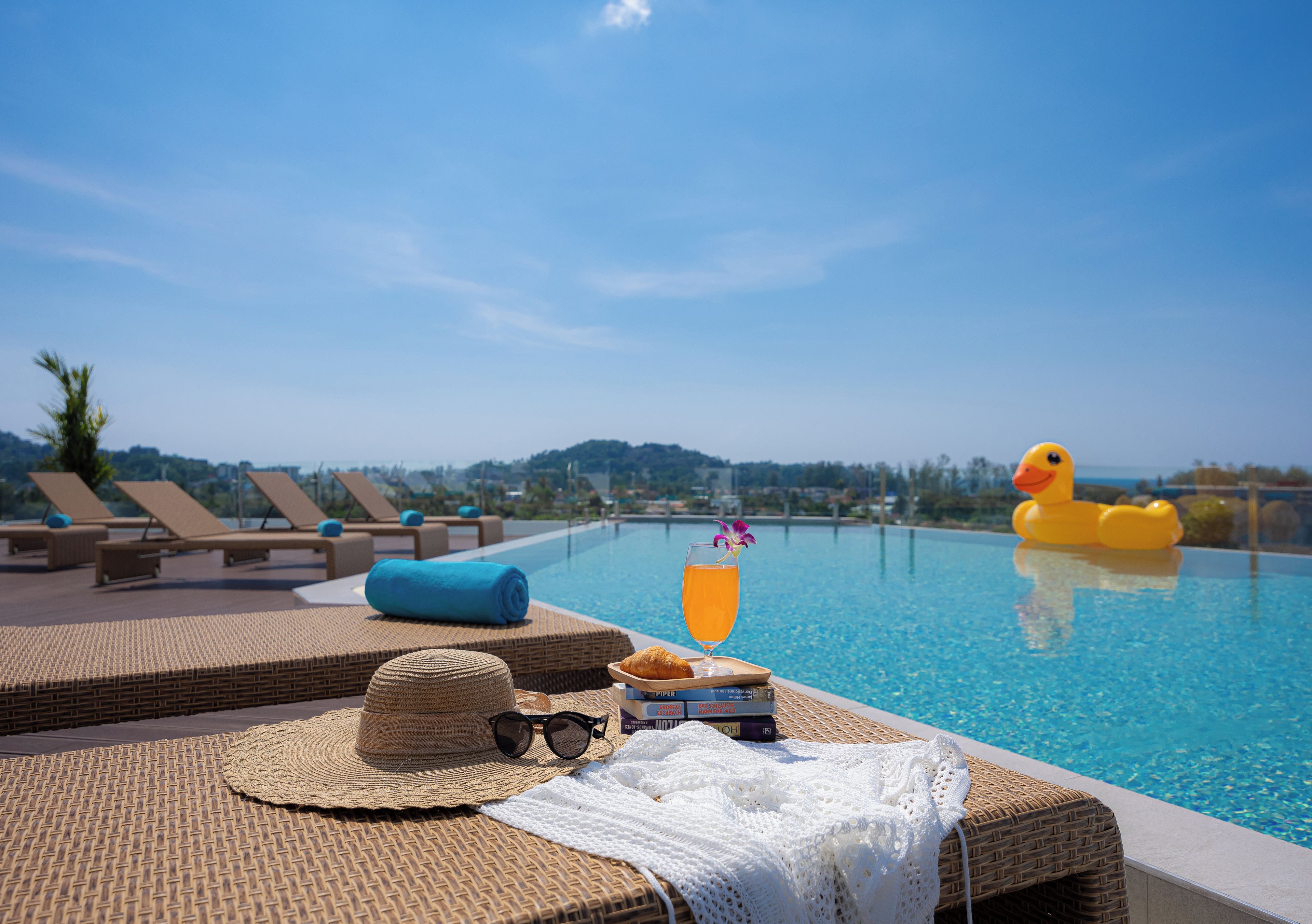 Escena junto a la piscina con sillas de descanso, un sombrero, gafas de sol, una bebida y un flotador con forma de pato.