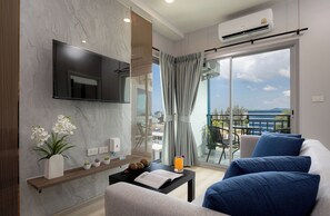 A modern living room with a flat-screen TV, a sofa with blue cushions, a coffee table with a book and a glass of orange juice, and a balcony with a view of the sea. - Skyline Bangtao Beach by RESAVA (Phuket)