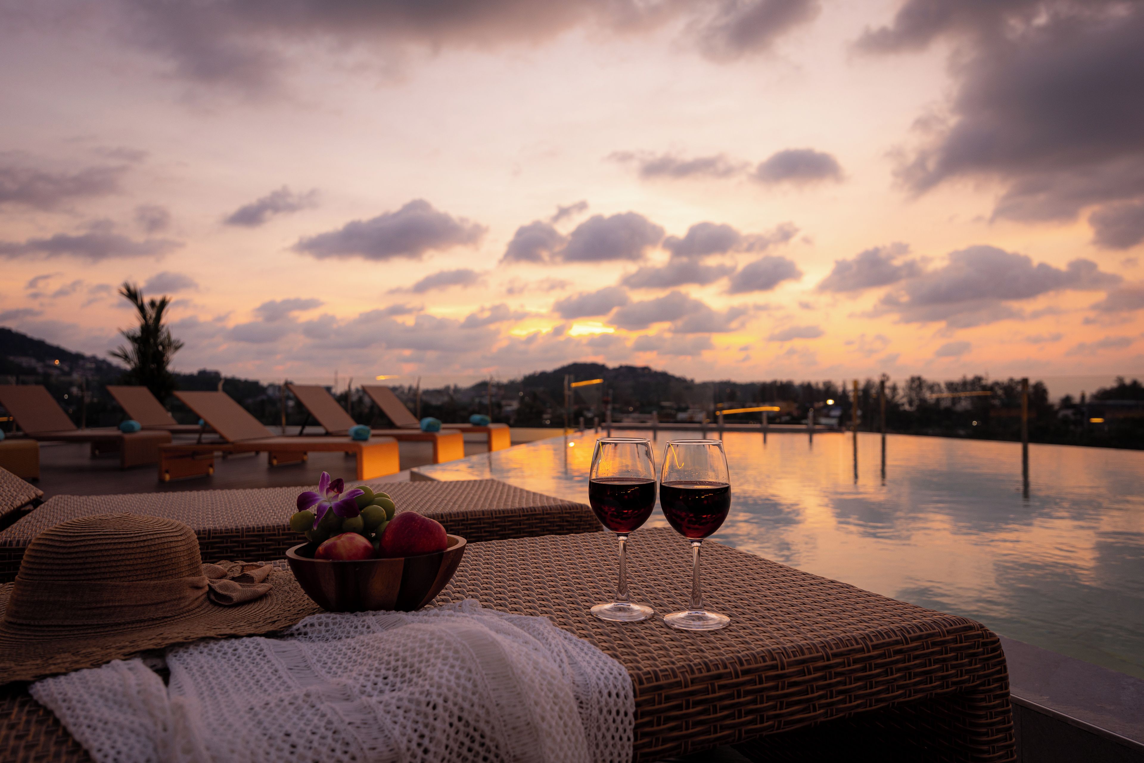 Une vue sur le coucher de soleil depuis un salon de bord de piscine, avec deux verres de vin rouge et un saladier de fruits.
