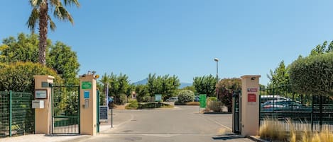 Une entrée fermée par un portail donnant sur une propriété, avec un ciel dégagé, un palmier et un espace de stationnement.