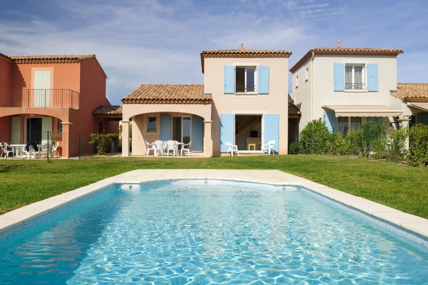 A swimming pool with a clear blue surface, surrounded by a grassy area and adjacent buildings with balconies and outdoor seating.