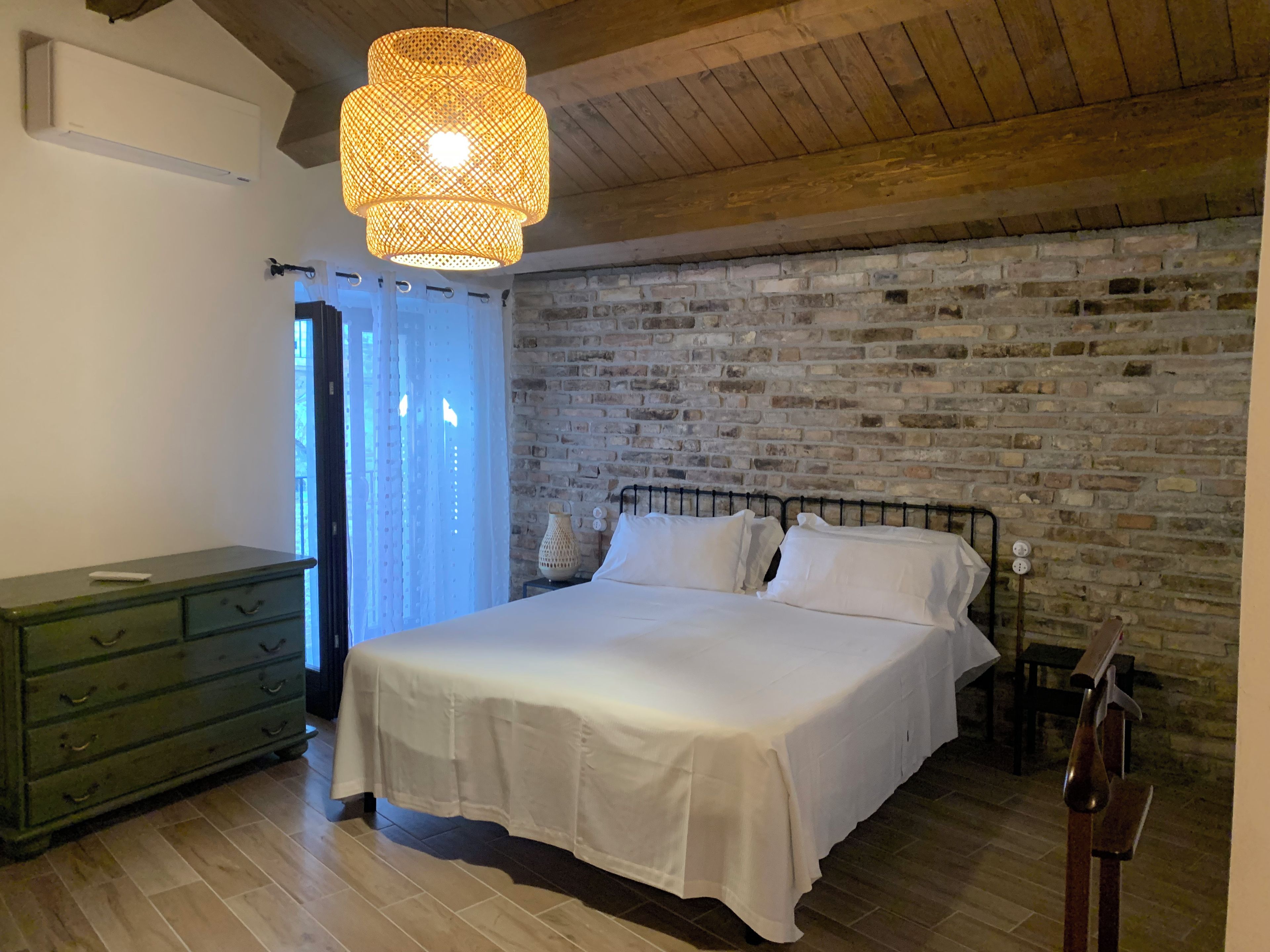A bedroom with a brick wall, a bed with white bedding, a green dresser, and a hanging light fixture.