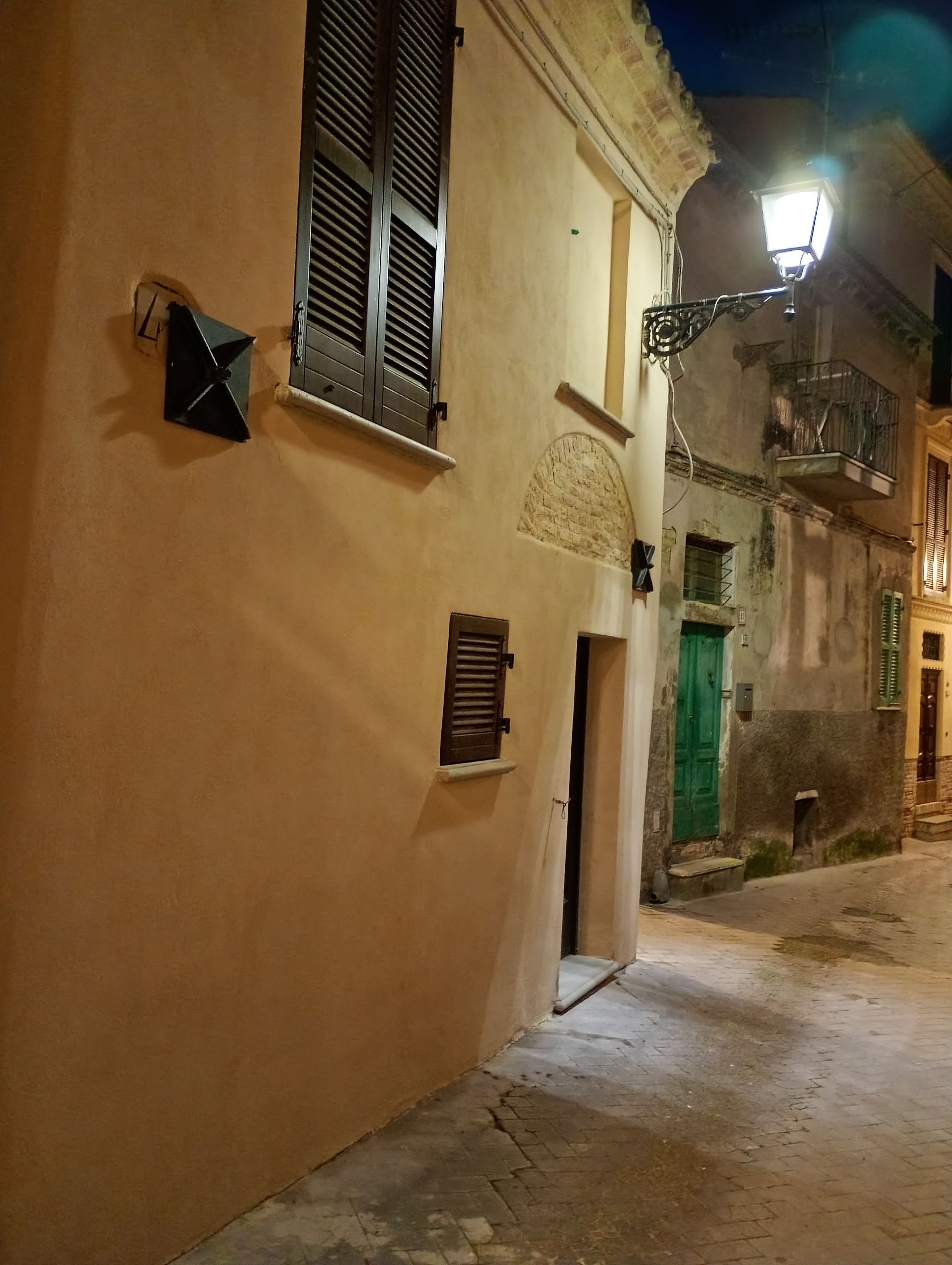 A narrow street with buildings on either side, a street lamp, and a door with a green shutter.