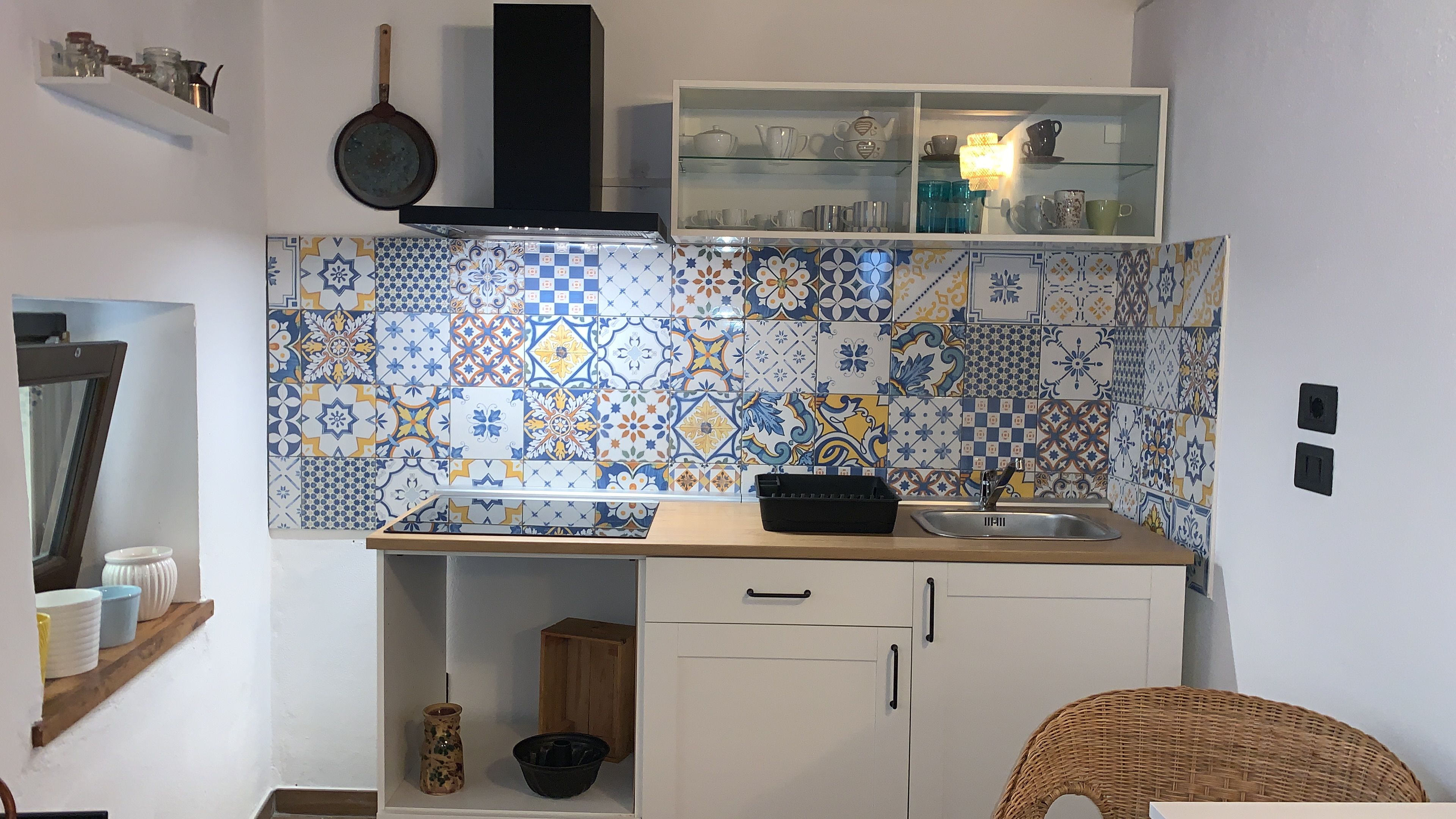 A kitchen with a tiled backsplash, a black range hood, a white cabinet with a glass shelf above, and a wicker chair.