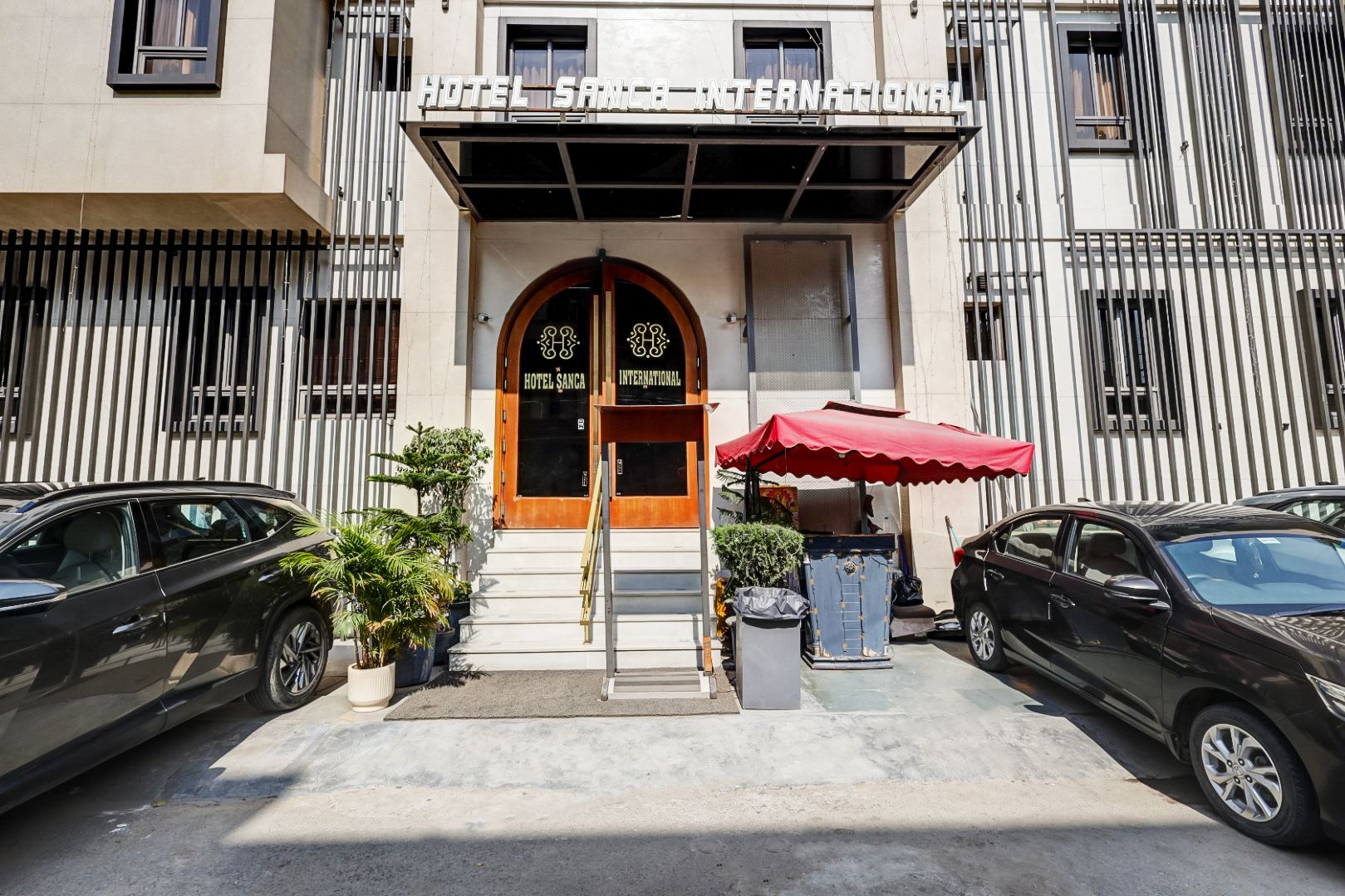 A hotel entrance with a double door, a red umbrella, and two cars parked in front.