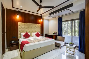 A hotel room with a large bed, a ceiling fan, a desk with a chair, and a window with curtains. - Sanca International Patel Nagar New Delhi (New Delhi)
