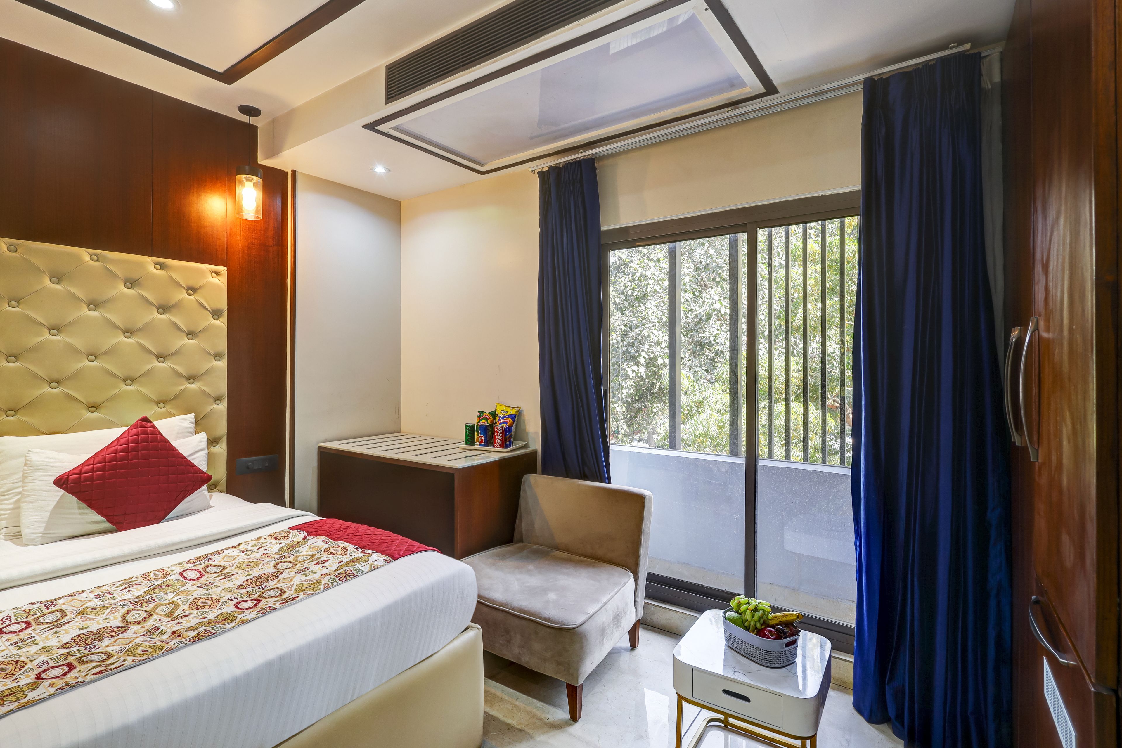 Habitación de hotel con cama, una mesita, una silla, ventana con cortinas y vistas a la vegetación.