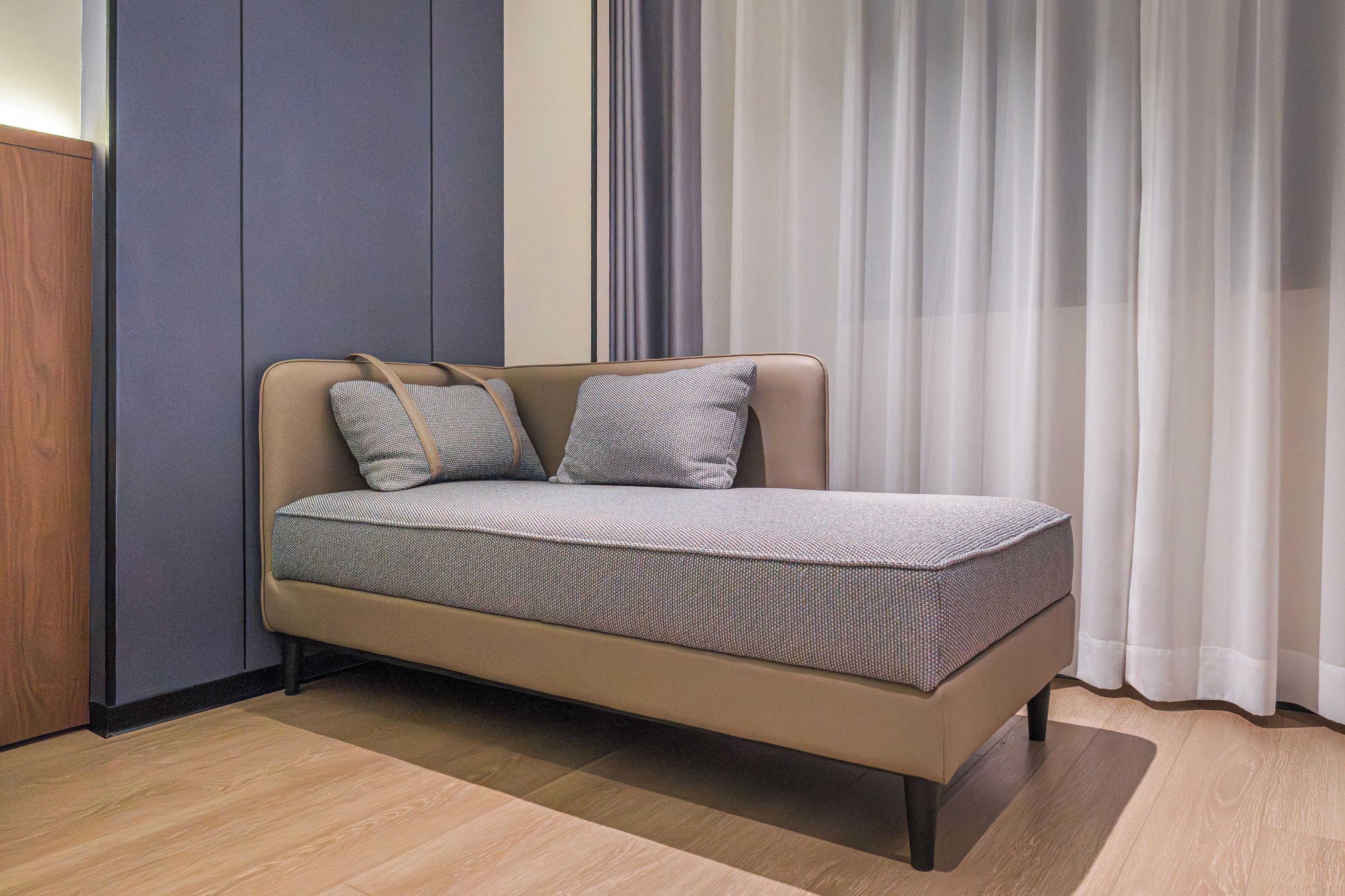 A single bed with a patterned bedspread and two pillows, positioned against a wall with a vertical panel and a window with sheer curtains.