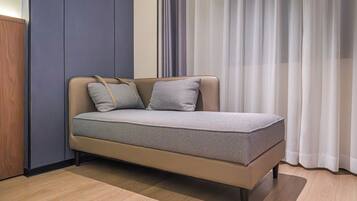 A single bed with a patterned bedspread and two pillows, positioned against a wall with a vertical panel and a window with sheer curtains.