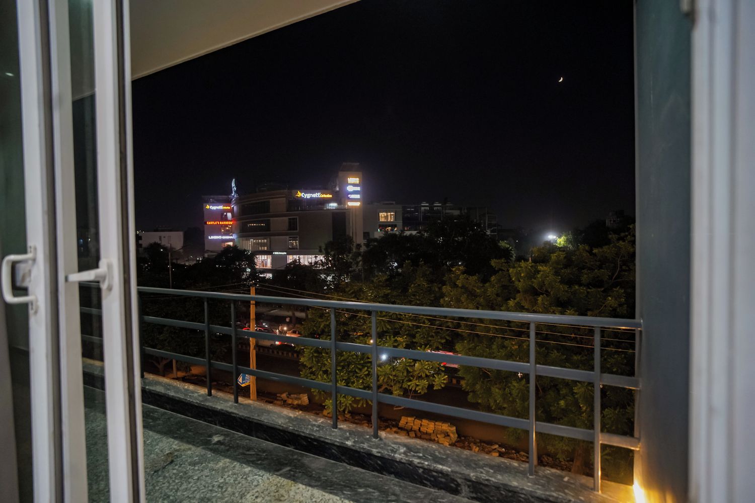 Vista nocturna desde un balcón con barandilla, con vistas a un paisaje urbano de edificios iluminados y árboles.