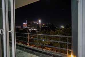 Vista nocturna des d'un balcó amb barana, amb una ciutat il·luminada i arbres a la vista.